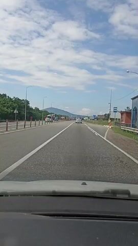 A forward-facing view from a vehicle on a paved highway. The highway has two lanes in each direction separated by a white line. Guardrails are present on the left side of the road, with trees and a mountain range in the background. Streetlights line the highway. Several vehicles, including trucks and cars, are visible on the road, some in traffic. The sky is blue with scattered clouds. The time stamps indicate the video is approximately 5 seconds long.