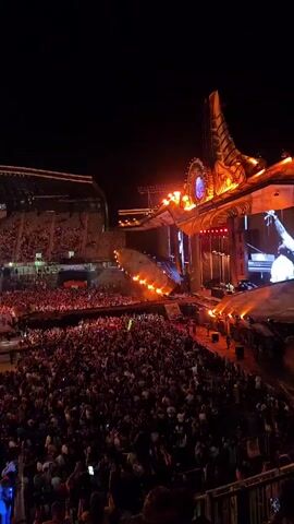The video is a first-person perspective of a large crowd gathered in a stadium at night for an event. The stage is visible in the distance, illuminated with bright lights. The audience is densely packed, with many people holding up their phones, likely recording or taking pictures. The overall atmosphere appears to be energetic and exciting, with the lighting and crowd suggesting a concert or similar large-scale performance.