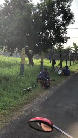 Two individuals are seated on the grassy verge beside a paved road. To the left, a person wearing a hat, sunglasses, and a blue jacket is crouched down, appearing to be tending to their footwear or belongings. Next to them, a grass trimmer lies on the ground. To the right, a second person, wearing a blue shirt, is also seated, possibly resting or eating from a bag. In the background, a fence, trees, and vegetation are visible. The overall lighting suggests it is daytime, possibly morning or late afternoon.