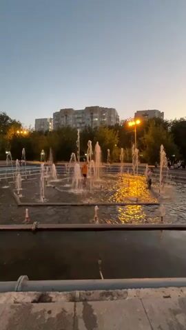 The video shows a fountain in a park at dusk. The fountain has multiple jets of water spraying upwards. There are a few people in and around the fountain. In the background, there are trees and buildings. The sky is a gradient of blue and orange, indicating the sun is setting. The water in the fountain is illuminated by artificial lights, creating a golden reflection on the surface.