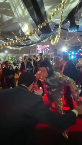 The video shows a party or event with a dance floor and a DJ booth. People are dressed in formal attire, suits and dresses. In the center of the dance floor, a person in a full monkey costume, covered in shimmering sequins, is dancing. The costume has LED lights on the legs that change color from red to blue. A person in a dark suit is facing the monkey character, with their back to the camera, and appears to be interacting with them, possibly dancing. Other guests are visible in the background, watching the performance and mingling. The ceiling is decorated with draped fabric in white and black, and gold bead garlands. Red and blue lights illuminate the dance floor. A screen in the background displays images or videos.