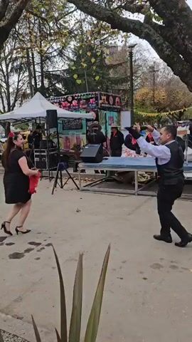 A man and woman are dancing on a concrete plaza. The woman is in a black dress and heels, holding a red fan. The man is in a black vest and slacks. They are dancing near a food stand with a tent. A band is playing music in the background. There are trees around the plaza.