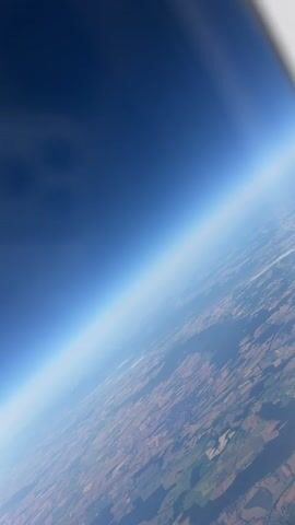The video captures a view from an airplane window, looking down at the earth from a high altitude. The horizon is clearly visible, with the dark blue of the upper atmosphere transitioning to a lighter blue and then to a hazy sky near the earth's surface. Below, a patchwork of fields and landforms is visible, suggesting a rural or agricultural landscape. The sun appears to be low on the horizon or just below it, casting long shadows and illuminating the scene with a warm, soft light. The edge of the airplane window is visible in the foreground, framing the view. There are no people or other objects clearly discernible in the video. The overall impression is one of vastness and perspective, highlighting the scale of the earth from above.