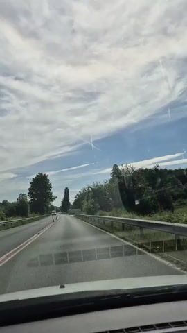 The video shows a view from inside a vehicle traveling on a paved road. The road has a double yellow line and a red line on the left side. On both sides of the road, there are guardrails and trees. The sky is partly cloudy with some streaks of cirrus clouds and a contrail from an aircraft. There are other vehicles on the road, including a white van in front of the camera vehicle and a white truck further ahead. The scenery is green and appears to be in a rural or suburban area. The video is taken during the daytime, with bright sunlight.