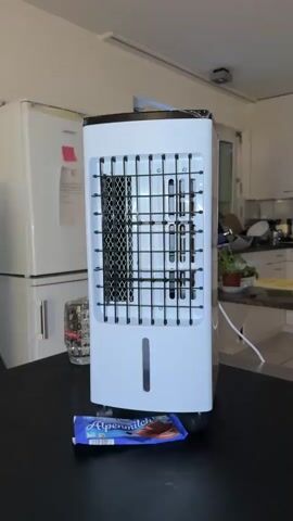 A close-up, stationary, eye-level shot captures the back of a white portable air cooler placed on a dark table. In front of the air cooler, on the table, is a wrapper for an "Alpenmilch" chocolate bar. The air cooler has a black grate on its upper half and a clear plastic water level indicator on its lower half. To the left of the air cooler, a glass filled with ice and a straw is visible. The background reveals a kitchen setting with a white refrigerator, countertops, cabinets, and a potted plant. The lighting suggests an indoor environment.
