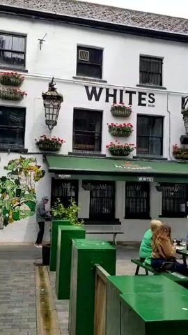 The video shows an outdoor seating area of a pub called "WHITES 1830". Several people are seated at green picnic tables, some of whom are drinking and conversing. A man in a blue jacket stands near the entrance to the pub, looking at his phone. Another person, in a purple jacket, walks away from the pub. The pub has a white facade with black-framed windows and green awnings. Decorative hanging lanterns are mounted on the building. Green planters filled with red flowers are attached to the wall above the awnings. The ground appears to be paved with cobblestones. The overall atmosphere is relaxed and casual.