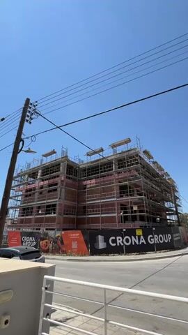 This is a vertical, outdoor, daytime video capturing a multi-story building under construction. The building is wrapped in scaffolding and appears to be made of brick with pink insulation visible. There is a black banner at the bottom of the frame with "CRONA GROUP" written in white text. To the left of the banner, there is a smaller orange banner with a QR code and what appears to be a phone number and website. The sky is clear and blue, suggesting fair weather. Power lines are visible crisscrossing the top of the frame, and a utility pole is on the left side of the image. The scene appears to be a street-level view of a construction site, likely in a developed area given the presence of overhead power lines and a paved road. No people are clearly visible. The overall impression is one of ongoing development and progress.