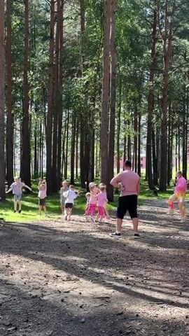 A group of children and one adult are in a wooded area. The adult, a man wearing a pink shirt and black shorts, stands facing away from the camera, looking towards a group of children. The children, varying in age, are spread out in front of him. Some are jumping, one is in mid-air doing a cartwheel, and others are standing or running. One child, a girl in a bright pink outfit, stands to the far right, also looking towards the group and appears to be dancing or participating in an activity. The ground is covered in dirt and fallen leaves, and tall pine trees surround the area, with sunlight filtering through the branches. The overall impression is of a playful outdoor activity or lesson.