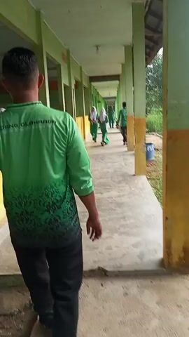 A group of people are walking down a covered outdoor walkway. The walkway has a concrete floor and is lined with green columns supporting a roof. On the left, there are rooms with yellow walls and open doorways, with some people seated inside. On the right, there is an outdoor area with grass and trees. Most of the people visible are wearing green uniforms, some with white hijabs. One person in the foreground is walking away from the camera, wearing a green shirt with black lettering that appears to say "OLAHRAGA" and black pants. The lighting suggests it is daytime.