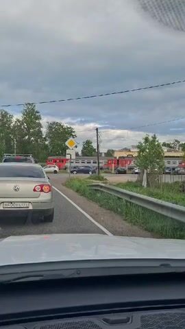 The video shows a view from the inside of a car, looking out at a road intersection in what appears to be a suburban or rural area. The car is stopped in traffic behind a silver Volkswagen.

On the right side of the road, a gray guardrail runs along a patch of grass. Beyond that is a small tree.  Further in the background, there's a building, partially obscured by the tree, and a train with red carriages parked behind it. There are several cars parked in front of the building as well. A yellow diamond-shaped sign is visible to the left of the building.

The sky is overcast with gray clouds. Power lines crisscross overhead.

The camera remains static for the duration of the video.