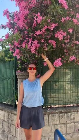 A young woman stands in front of a blooming pink oleander tree and a green fence. She is wearing a light blue tank top, dark gray shorts, and sunglasses. She has her right arm raised, touching the flowers. The woman is smiling and looking at the camera. The background is a sunny day with clear blue sky.