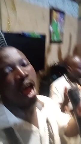 The video begins with a close-up of a Black man in the foreground, speaking into a microphone with his mouth open. He is wearing a light-colored collared shirt and multiple strands of beaded necklaces. In the background, another Black man is visible, also wearing a light-colored shirt and raising his arm. A framed picture hangs on the wall behind them, and a fan is partially visible on the left. The environment appears to be indoors, with a television or screen visible on the left side of the frame. The camera angle is low, looking up at the man with the microphone.