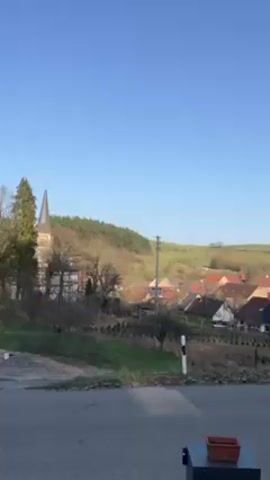The video presents a daytime view of a small European village nestled in a valley. In the foreground, a paved road stretches horizontally across the frame, with a small, dark, boxy object containing a clay-colored planter sitting on its right side. Beyond the road, the landscape rises gently, dotted with houses featuring predominantly red-tiled roofs. A church steeple is visible on the left, amidst bare trees. A forest covers the hillside in the background. The sky is a clear, cloudless blue. The overall lighting suggests a bright, sunny day, enhancing the natural colors of the scene.