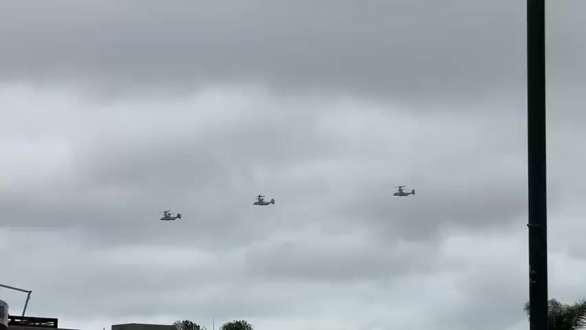 Three Bell Boeing V-22 Osprey tiltrotor aircraft fly in formation across a cloudy sky. The aircraft are spaced evenly apart, with the lead aircraft slightly to the left and lower than the other two. In the foreground, the top of a green and white bus with "TROLLEYTOURS.COM" written on it is visible. A lamppost is also visible on top of the bus. The overall impression is one of a military flyover in an urban or suburban environment.