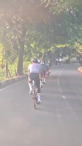 This video shows a group of cyclists riding down a paved road lined with trees. The sun is low in the sky, casting long shadows, suggesting it's either early morning or late afternoon. The lighting is warm and the trees are lush and green, indicating a pleasant, possibly tropical climate. The cyclists are wearing helmets and athletic clothing, with one rider in the foreground sporting white socks and black shorts. They appear to be engaged in a cycling group ride or training session, moving in the same direction. A red truck is visible on the right side of the road, also moving in the same direction. The road curves to the right, with a low concrete barrier on the left side. The overall mood is active and perhaps focused, given the cycling activity. There is some visible text on the side of the red truck, but it is too blurry to be legible in the provided frames. The location is identified as Santan, Indonesia.
