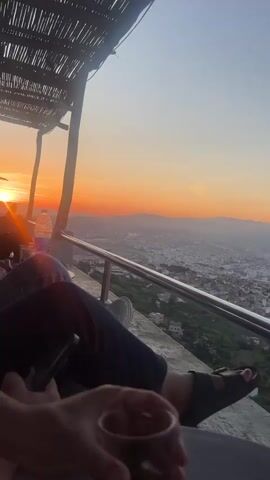 The video captures a serene sunset scene from a high vantage point overlooking the city of Tétouan, Morocco. The sky is painted with vibrant orange, yellow, and light blue hues as the sun dips below the horizon, casting long shadows and a warm glow over the landscape. In the foreground, a person's legs and feet are visible, suggesting they are seated and relaxed, enjoying the view. One hand holds a smartphone, while another reaches for a clear glass containing a drink, possibly mint tea, a common beverage in Morocco. A plastic water bottle is also visible nearby.

The setting appears to be an outdoor cafe or viewpoint, characterized by a rustic structure with wooden beams and a slatted, possibly thatched or reed, roof. A metal railing extends along the edge, providing a safety barrier and a clear line of sight to the sprawling city below. The cityscape stretches into the distance, with buildings covering the undulating terrain, and mountains forming a hazy backdrop on the horizon. The overall atmosphere is one of tranquility and enjoyment of the scenic vista.