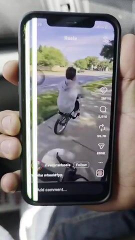 A person is holding a black smartphone vertically, displaying a video on its screen. The phone's screen has noticeable damage, with several bright green vertical lines running down the left side.

The video playing on the screen is a "Reel," likely from a social media platform like Instagram or Facebook. It shows an outdoor scene with a person riding a bicycle on what appears to be a paved path or sidewalk next to a grassy area with trees. The biker, wearing a light-colored hoodie and dark shorts, is attempting a maneuver, possibly a wheelie or a jump. The camera perspective in the video seems to be from another bike or a low angle, following the rider.

Visible text and icons on the smartphone screen include: "Reels" at the top, a user handle "riveronwheels" with a "Follow" button, and partially visible hashtags "bike wheel#fyp". Interaction statistics for the video are also shown: "2M" likes (heart icon), "5,515" comments (speech bubble icon), "65.7K" shares (paper plane icon), and "850K" saves (bookmark icon). Below the video, there's an "Add comment..." prompt. The top status bar of the phone shows the time "1:29", signal strength, "5G+", and battery level.

The individual holding the phone has light brown skin, and only their thumb and fingers are clearly visible. The background behind the phone is blurred, suggesting the person is inside a vehicle, possibly a car, given the reflections and texture of what looks like an interior. The time of day appears to be daytime, with bright ambient light in the video.