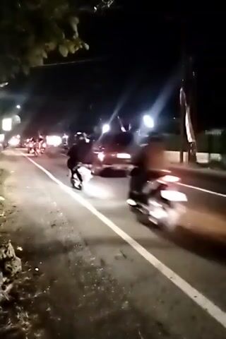A video captures a nighttime scene on a road with multiple vehicles. In the foreground, a dark-colored SUV is on the right side of the road, its taillights illuminated. To the left of the SUV, a white pickup truck travels in the same direction. Further ahead on the road, several motorcycles are visible, some with riders. Streetlights and the headlights of vehicles illuminate the dark environment. The road appears to have a white dividing line. The camera is positioned at a low angle, likely from another vehicle or a person on the side of the road, as evidenced by the perspective and movement. The overall impression is of a convoy or group of vehicles moving together at night.