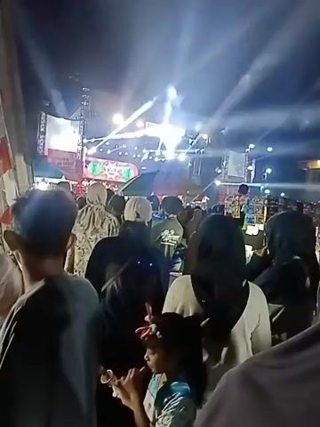 The video shows a nighttime outdoor concert or event. A crowd of people are gathered, facing a brightly lit stage. The stage features a large screen or backdrop, multiple lights, and a truss system. Beams of white light are cutting through the darkness from the stage. The audience members are mostly obscured by darkness and the angle of the shot, but many appear to be wearing casual clothing and some have head coverings. The atmosphere appears to be festive and energetic, with the lighting suggesting a performance is underway.