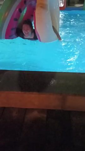 A person is partially submerged in a bright blue swimming pool, wearing a black shirt. They are holding onto an inflatable toy that appears to be a large ring with a watermelon design on one side and a solid white color on the other. The person's face is visible, and they are smiling. The camera is positioned above the water, looking down at the person. The edge of the pool, made of wood and dark stone or tile, is visible in the foreground. The lighting suggests it is nighttime or indoors, as there are artificial lights illuminating the pool and the surrounding area.
