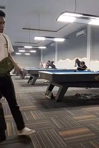 A medium shot shows a group of people playing billiards. A young man in a white t-shirt and black pants stands in the foreground, facing left. He has a green messenger bag slung across his body. In the background, three men are playing billiards at separate tables. The room is lit with fluorescent lights, and the walls are painted gray with a mural of mountains. The floor is carpeted in a geometric pattern of browns and grays.