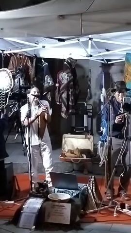 A man in white pants and a white shirt stands in front of a stall, playing a pan flute and singing. He is illuminated by a white tent with lights. Behind him, various items are displayed, including blankets and baskets. To his left, another man in a dark jacket and baseball cap holds a camera on a tripod, filming the performance. A laptop sits on a table in front of the flute player, along with papers and a plate. The scene appears to be outdoors at night.