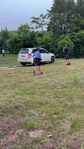 Two individuals are in a grassy area with a white SUV parked in the background. The person in the foreground, a younger individual wearing a purple shirt and black shorts, runs towards the right of the frame. The second person, an adult male wearing a dark shirt and red shorts, stands near the SUV, looking towards the younger individual. The environment appears to be a rural or semi-rural setting with trees and vegetation surrounding the open grassy area. The sky is overcast, suggesting cloudy weather.