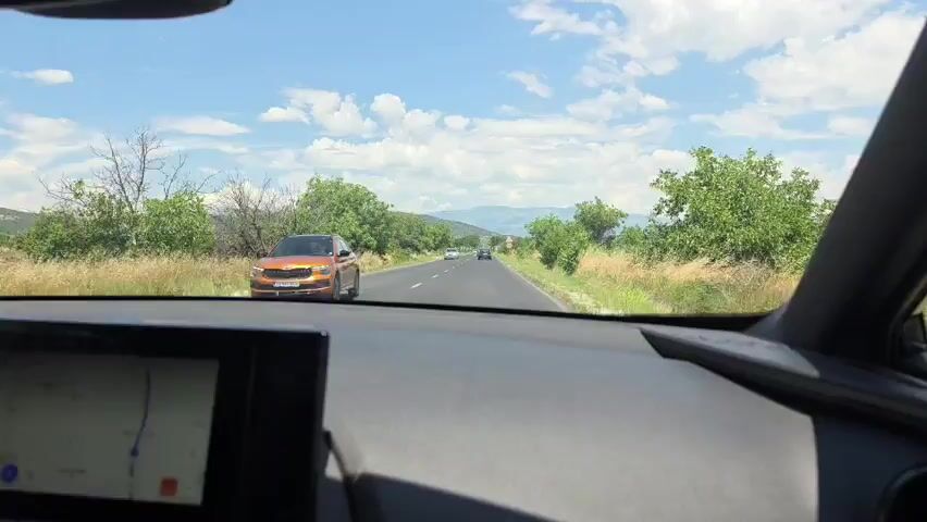 The video shows a first-person perspective from inside a car driving on a paved road. The dashboard with a navigation screen is visible in the foreground. The road stretches ahead through a rural landscape with green trees and vegetation on either side, under a bright blue sky with scattered clouds. Distant hills or mountains can be seen in the background. There are other vehicles on the road ahead. A speed limit sign is visible on the right side of the road.
