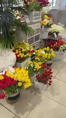 The video shows a room in a store with a variety of artificial flowers, including tulips and calla lilies, arranged in baskets and containers. The tulips are primarily red, yellow, and white. Some of the flower arrangements are displayed on small wooden tables. Behind the flowers is a decorative chest of drawers and a cashier's counter with a female employee. The store shelves in the background display various household goods. Crying or fussing noises can be heard, possibly from a baby or child.