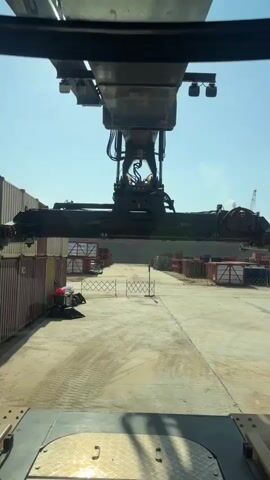 The video shows a view from inside a crane cab, looking forward and slightly down. A large crane spreader bar is in the foreground, suspended from a larger overhead crane structure. The spreader bar is positioned above a row of shipping containers. The environment appears to be a shipping yard or port, with more containers visible in the background and a clear blue sky overhead. There are no people or direct interactions visible in the footage, only the movement of the crane and its spreader bar as it likely prepares to lift or move a container. The overall scene depicts industrial logistics in operation.