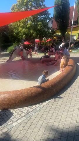 The video shows a shallow splash pad area with several children playing in the water. A large, brown, textured structure, resembling a snake, encircles a portion of the splash pad. In the background, several adults are visible, some sitting on benches and others supervising children. A red shade sail is stretched over the area, providing shade. The ground is paved with interlocking bricks. The environment appears to be an outdoor public space, likely a park or a water playground, on a sunny day.