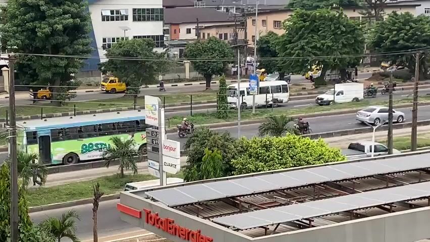 The video captures a street scene from an elevated perspective, likely from a building overlooking a busy road. In the foreground, a TotalEnergies gas station canopy is visible, covered with solar panels. The road itself features multiple lanes of traffic, with cars, buses, vans, and motorcycles navigating the street. Several vehicles are in motion, including a white van and a blue bus on the main road. On the sidewalk, there are signs, trees, and some greenery. In the background, buildings of various sizes and architectural styles line the street, interspersed with more trees. The overall environment appears to be an urban setting with moderate traffic and infrastructure typical of a city. The lighting suggests it is daytime, possibly overcast or in the afternoon. There are no discernible interactions between people, and the focus is on the movement of vehicles and the general urban landscape.