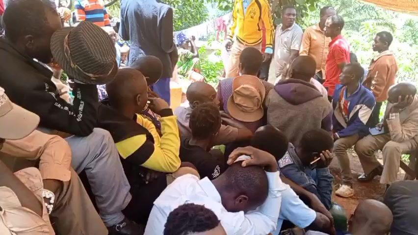 A group of about 25 men, women, and children are gathered under a makeshift tarp awning in a partially shaded outdoor area. Most of the group is seated on the ground or on wooden benches, while some individuals stand. The people are wearing casual clothing, with some men in collared shirts and jackets. A man in a yellow jacket is gesturing with his hand and speaking to a man in a suit. The scene appears to be a gathering or meeting of some kind. The surroundings include greenery, trees, and a partially visible building or structure.