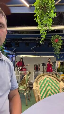 The video captures a live music performance at an outdoor venue at night. In the foreground, a man is partially visible, looking towards the camera. In the background, on a raised stage, two performers are actively engaged in their music. One individual, wearing sunglasses and a red shirt, is playing a red electric guitar. The other performer, a woman in a red dress, is singing into a microphone. Two large fans are positioned behind the performers. The stage is illuminated by overhead lights. The seating area in front of the stage is furnished with wicker chairs with green and white patterned cushions, and tables covered with white tablecloths. Lush green plants hang from above, adding a natural element to the environment. The overall scene suggests a relaxed evening entertainment setting.
