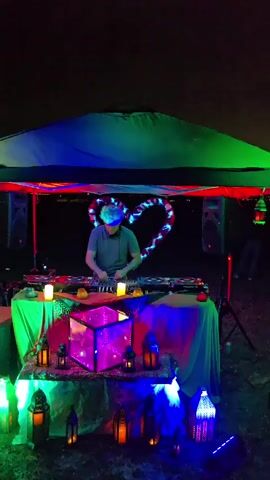 A person is DJing under a green tent at night. They are wearing a grey shirt and have headphones on. There are colorful lights illuminating the area, including a heart-shaped light behind the DJ. A cubic structure with colorful lights sits on the table in front of the DJ. Several lanterns are hung around the tent, emitting a warm glow. In the background, two other people are visible, one standing and the other seated. The sky above is dark with some clouds. A small, illuminated object, possibly a drone or a kite, is visible in the distance, moving across the sky.