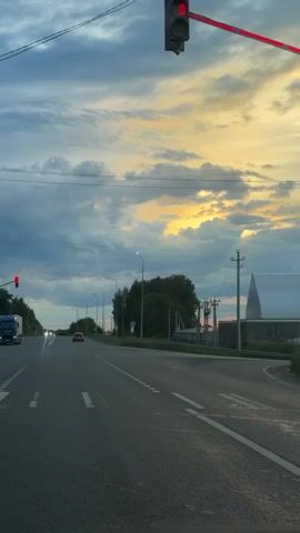 This video captures a view from a vehicle driving on a road, likely in Russia based on the context. The scene is set during what appears to be a cloudy sunset, with hues of orange and yellow visible through breaks in the dark grey clouds.

The primary subjects are the road and the sky. The road is paved and marked with white lines. In the distance, vehicles, including a truck and cars, can be seen on the road. There are utility poles and streetlights lining the road, suggesting an urban or suburban setting. Trees are visible on the right side of the road. A distinct, tall, cone-shaped structure stands out against the sky on the right, near some buildings.

The activity taking place is driving along the road. Traffic lights are visible and appear to be changing. One traffic light prominently displays an orange (amber) light at the beginning of the clip, and later changes to a green light with a digital countdown timer showing "41" seconds.

Notable details include the dramatic cloudy sky with a sunset glow, the presence of traffic control signals, and the unique cone-shaped structure. The overall mood conveyed is one of travel and the transition from day to night under a dynamic sky. There is no visible text in the video itself, other than the digital timer on the traffic light.