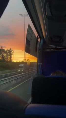 The video is a first-person perspective from inside a moving vehicle, likely a bus or train. The view is out the window on the left side, showing a sunset with orange and yellow hues. There are also train tracks visible. The interior of the vehicle is dark, with overhead lights and seats. A digital display is visible on the ceiling showing numbers and text in Cyrillic. The perspective shifts slightly within the vehicle, showing the backs of seats and some passengers.