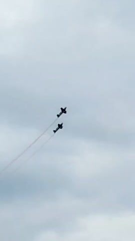Two dark-colored biplanes perform an aerial maneuver against a cloudy, overcast sky. The planes fly in close formation, trailing thin, reddish-orange smoke plumes that curve and dissipate as they descend. The planes appear to be angled upward and to the left, moving in a diagonal path. The camera angle is looking up at the aircraft.