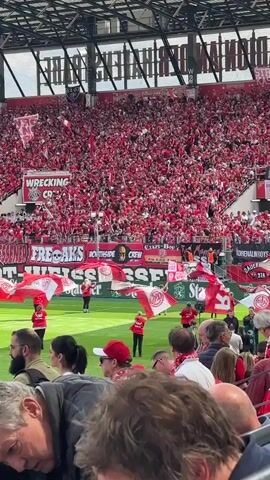 The video captures a crowded sports stadium filled with fans predominantly dressed in red. Several individuals in red uniforms stand on the green field holding and waving red and white flags. A large banner is displayed near the field with text in white and green. A variety of signs and banners with different text are visible above the fans. The fans are packed closely together and are waving their hands and flags. The stadium structure includes a metal roof. People in the foreground are watching the scene unfold.