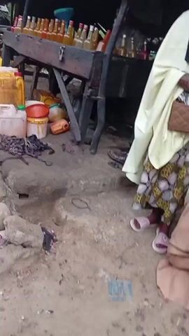 The video displays an unpaved outdoor market scene with multiple makeshift stalls featuring thatched or corrugated roofs supported by metal poles. Numerous yellow and white plastic jerrycans are on the ground. A wooden stand holds approximately two dozen clear glass bottles containing an amber liquid, many with red caps, alongside a blue funnel. A dark-colored motorcycle with a white bag strapped to its rear seat is parked on the dirt ground, which also shows red stains. A man in a light pink long robe and cap is seen adjusting the white bag on the motorcycle. Another man in a light brown long robe stands near the motorcycle, partially obscured. Other indistinct figures are visible in the background stalls. The camera pans across the scene.