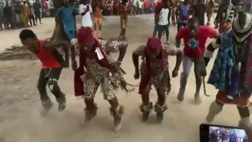 The video captures a vibrant cultural performance taking place outdoors on a sandy surface. A group of male dancers, dressed in an array of costumes, are the focal point. Some wear red tops and dark pants, while others are adorned in patterned garments resembling animal prints, paired with red headwear. One individual stands out with a more elaborate costume consisting of a patterned cloak, headwear resembling a mask, and other decorative elements. The dancers are engaged in synchronized movements, bending forward and seemingly stomping their feet, generating dust from the sandy ground. Spectators surround the performance area, observing the spectacle with interest. A mobile phone can be seen in the foreground, indicating that someone is recording the event. A tree is also present in the background, further establishing the outdoor setting. The overall atmosphere appears to be one of celebration and cultural expression.
