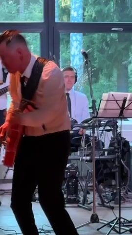 A man in a white collared shirt and black pants plays an orange electric guitar and sings into a microphone. He is standing on a wooden floor in front of a large window. Behind him, another man in a white shirt and black tie plays a bass guitar. Further behind, a person wearing headphones plays drums. The scene appears to be a live musical performance.