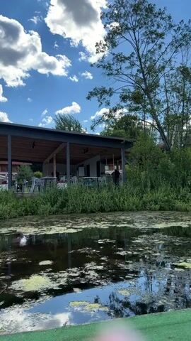 A video captures a scenic view of a body of water with aquatic plants, a green boat, and a covered outdoor seating area with tables and chairs. In the background, there is a red building with windows and trees. The sky is blue with scattered white clouds. A person in an orange vest can be seen near the seating area. Another boat is stacked on top of the green boat.