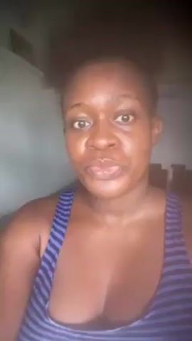 A woman with brown skin and dark hair tied up in a puff is centered in the frame, looking at the camera and smiling. She is wearing a blue and black striped tank top. The background is blurry but suggests a simple room. The lighting is natural.