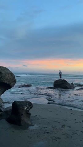 A person stands on a rock near the ocean, holding a fishing pole. The ocean has waves with white foam. The beach has dark sand and large dark rocks. The sky is a mix of blue, gray, pink, and orange. It appears to be either sunrise or sunset. The person is facing the ocean.