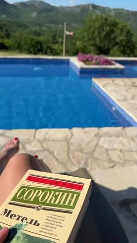 The video shows a person's legs, with red painted toenails, resting on a lounge chair next to a swimming pool. A book with Russian text on the cover, titled "SOROKIN" and "METEL," lies on the person's lap. The pool is bright blue and surrounded by stone paving. In the background, a lush green landscape of hills and trees stretches towards a clear blue sky. Another person, partially visible, is in the pool. The scene appears to be a sunny, outdoor vacation setting.