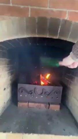 A person fans a fire in a brick fireplace with a green fly swatter. The fire burns in a decorative metal container sitting atop two bricks. Sparks fly from the fire, and the fireplace opening is dark. The person is wearing a brown plaid long sleeve shirt.