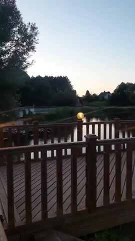 A wooden deck with a railing overlooks a calm body of water. Two illuminated spherical lamps are mounted on the railing posts. Trees and a house are visible in the background, silhouetted against a pale sky, suggesting it is dusk or dawn. The water reflects the sky and the lights on the railing.
