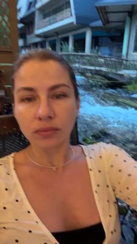 The media file features a woman in the foreground, facing the camera and appearing to be speaking or recording a video. She has light skin, light-colored eyes, and is wearing a white blouse with black polka dots over a black top. A thin necklace with a small pendant is visible around her neck.

The setting is outdoors, with a body of water, likely a river or stream, flowing in the background. The water is turbulent and appears to be moving rapidly, with rocks and vegetation visible along its banks. A rustic bridge with stone railings spans the water in the mid-ground. Beyond the bridge, there's a multi-story building with balconies and a neutral facade, suggesting it could be a hotel or a similar establishment. The overall ambiance suggests a natural setting with some man-made structures integrated into it.

The lighting appears to be low, indicating it might be dusk or evening. The woman's expression is neutral, and her gaze is directed towards the camera, suggesting an act of communication or recording. The camera seems to be positioned close to her, creating a sense of immediacy.

The overall scene suggests a leisurely moment, perhaps a traveler documenting their experience at a scenic location. The presence of the building and the bridge indicates a developed area, likely a tourist spot. The location context places this scene in Travnik, Bosnia and Herzegovina. There is no visible text in the media file.