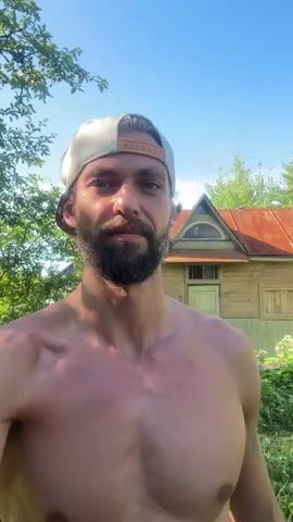 A man with a beard and wearing a baseball cap is shirtless and looking at the camera. He is standing outside in a natural setting with trees and a building in the background. The sky is blue and clear.